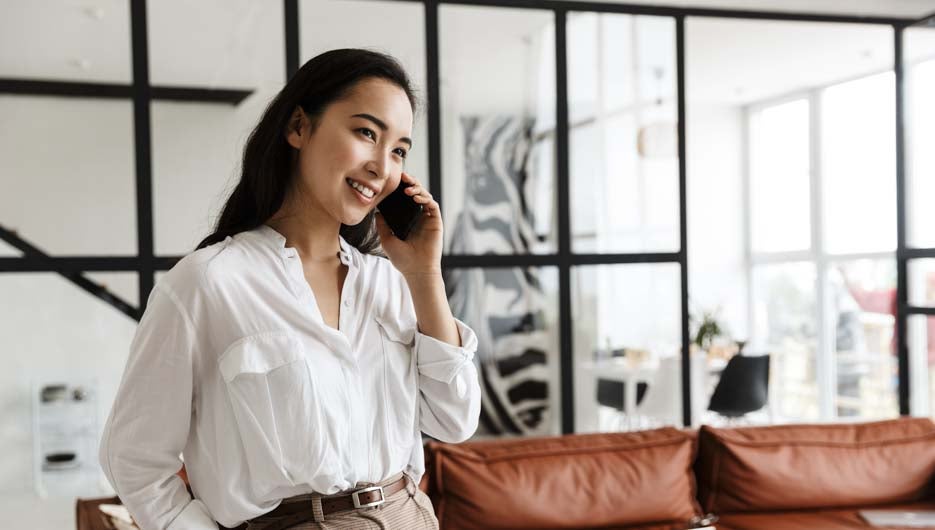 Beautiful smiling young asian woman wearing white shirt standing in a modern living room, talking on mobile phone