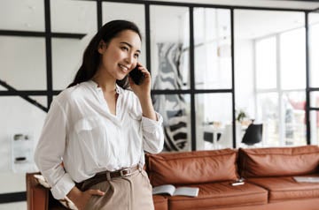 Beautiful smiling young asian woman wearing white shirt standing in a modern living room, talking on mobile phone